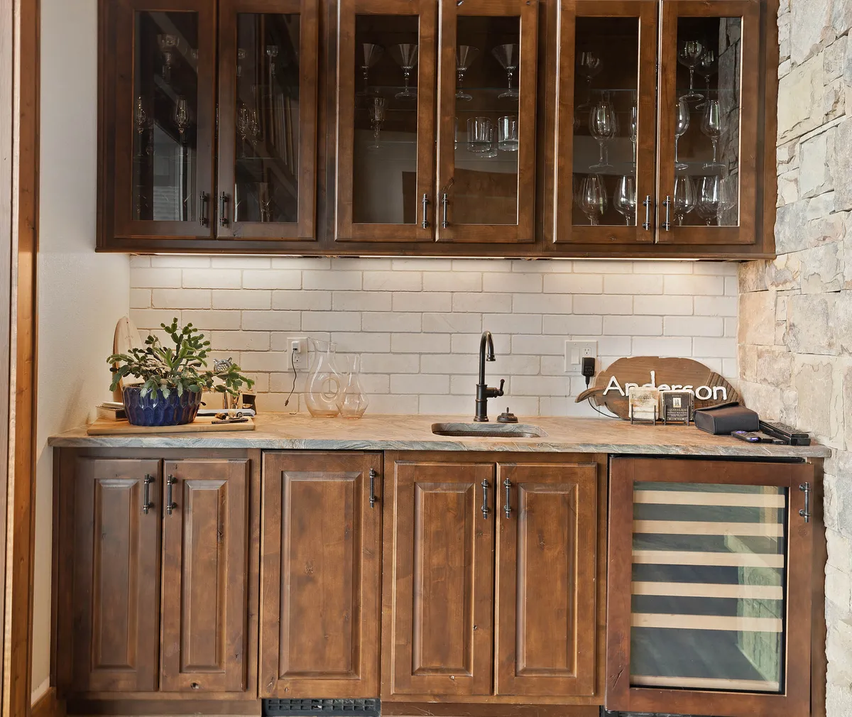 A stylish wet bar area with elegant wooden cabinetry, glassware on display, a wine cooler, and a sink. This functional and welcoming setup is ideal for preparing drinks and hosting guests, complementing the lounge environment.