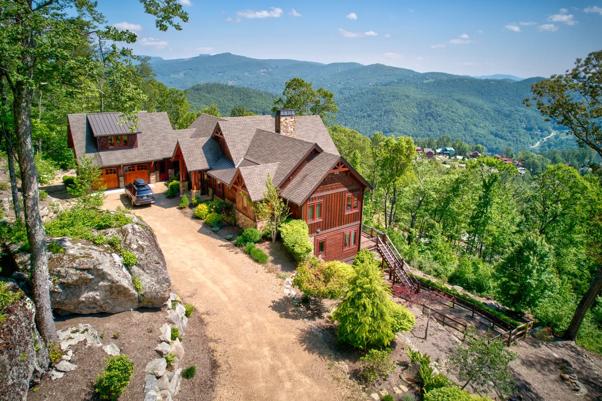 A welcoming exterior view of the large wooden house at Eagles Nest, featuring a spacious deck with outdoor seating, surrounded by lush trees and offering a distant mountain view. The scene is set up for relaxation and social gatherings, highlighting the connection to nature and the ample space for large groups or families.