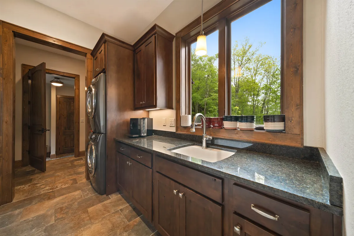A bright, open kitchen with granite countertops, elegant wooden cabinetry, and expansive windows allowing sunset views. The space is designed for group cooking and socializing, with a clear connection to the outdoors.