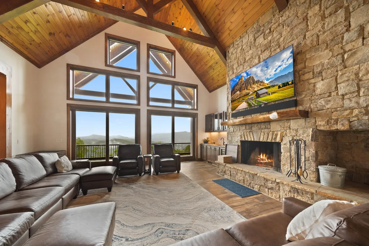 A spacious great room featuring a soaring stone fireplace, large sectional sofa, reclining chairs, and a large flat-screen TV. Expansive windows reveal sweeping mountain views, allowing natural light to flood the space. The room exudes comfort while offering a grand, open atmosphere perfect for gatherings.