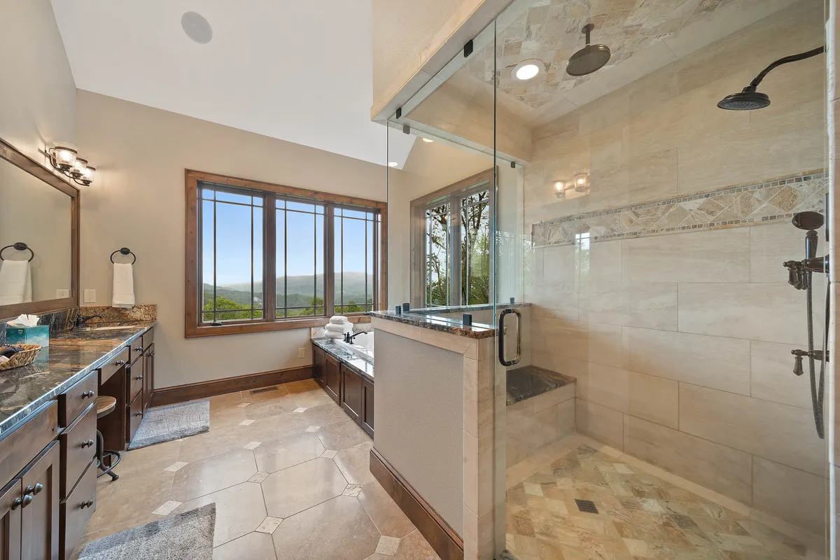 This luxurious bathroom features a freestanding bathtub placed beside expansive windows that offer sweeping mountain views. A double vanity with dark wood cabinetry and a spacious glass shower enclosure add to the spa-inspired ambiance, while natural light fills the space, creating a serene retreat.