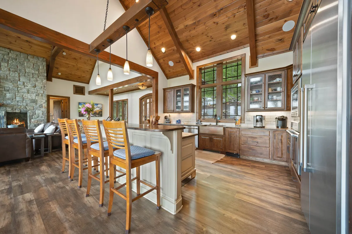 An inviting interior shot highlighting a beautifully designed kitchen with a large island, elegant wooden beams, bar stools, and a grand stone fireplace. The space is filled with warm, ambient light and features high-end appliances and finishes, suggesting opportunities for gourmet dining and cozy evenings by the fire.
