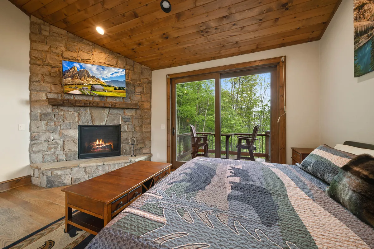 A tranquil primary bedroom suite featuring a plush king bed, a cozy stone fireplace, and large windows overlooking lush natural scenery. The room is accented by warm wood details, creating an inviting, restful atmosphere ideal for unwinding at the end of the day.