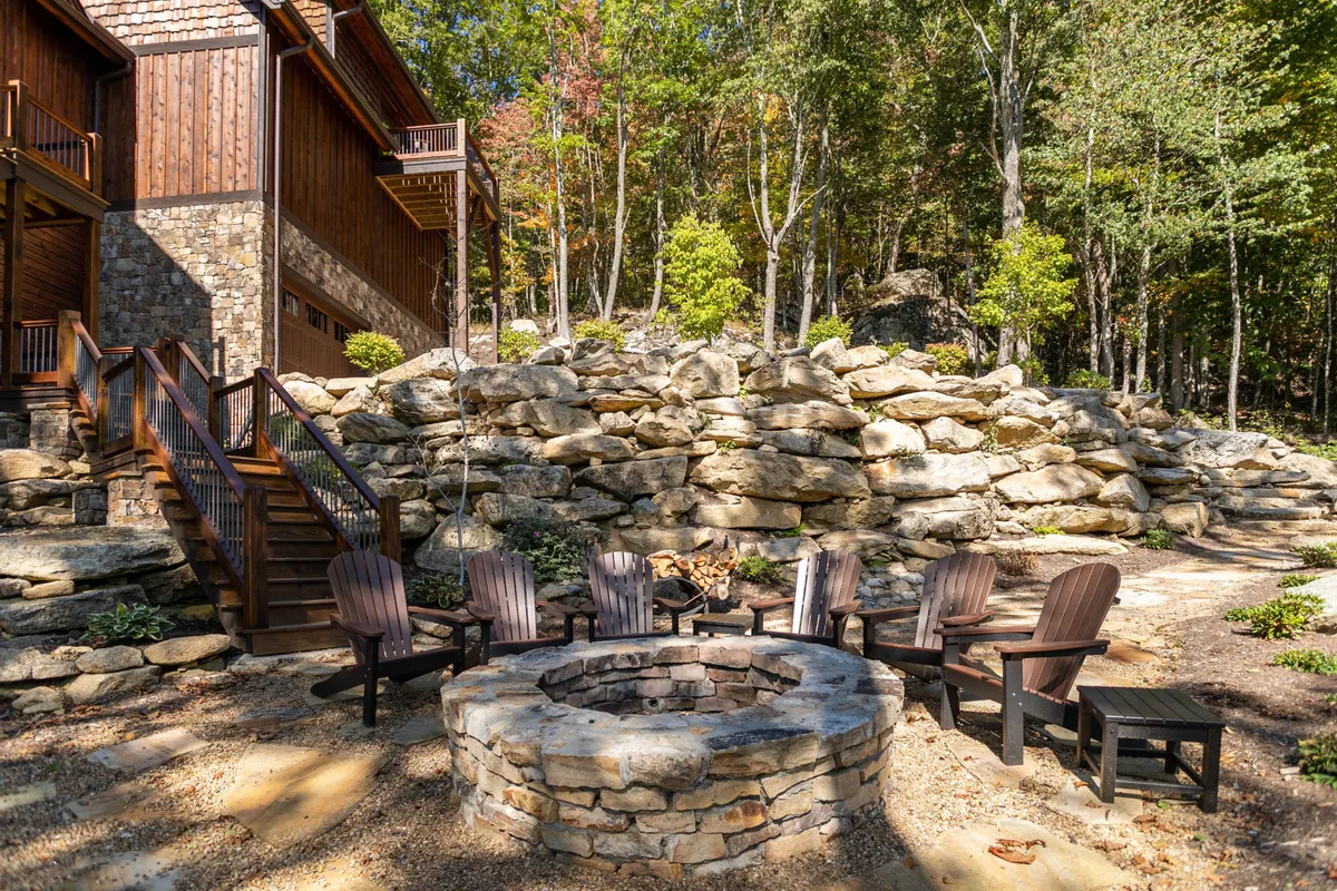 A cozy, outdoor fire pit area with wooden chairs arranged in a circle atop stone steps. The setting is surrounded by landscaped rocks and woodland, perfect for storytelling, toasting achievements, or simply enjoying the crisp mountain air under starry skies.