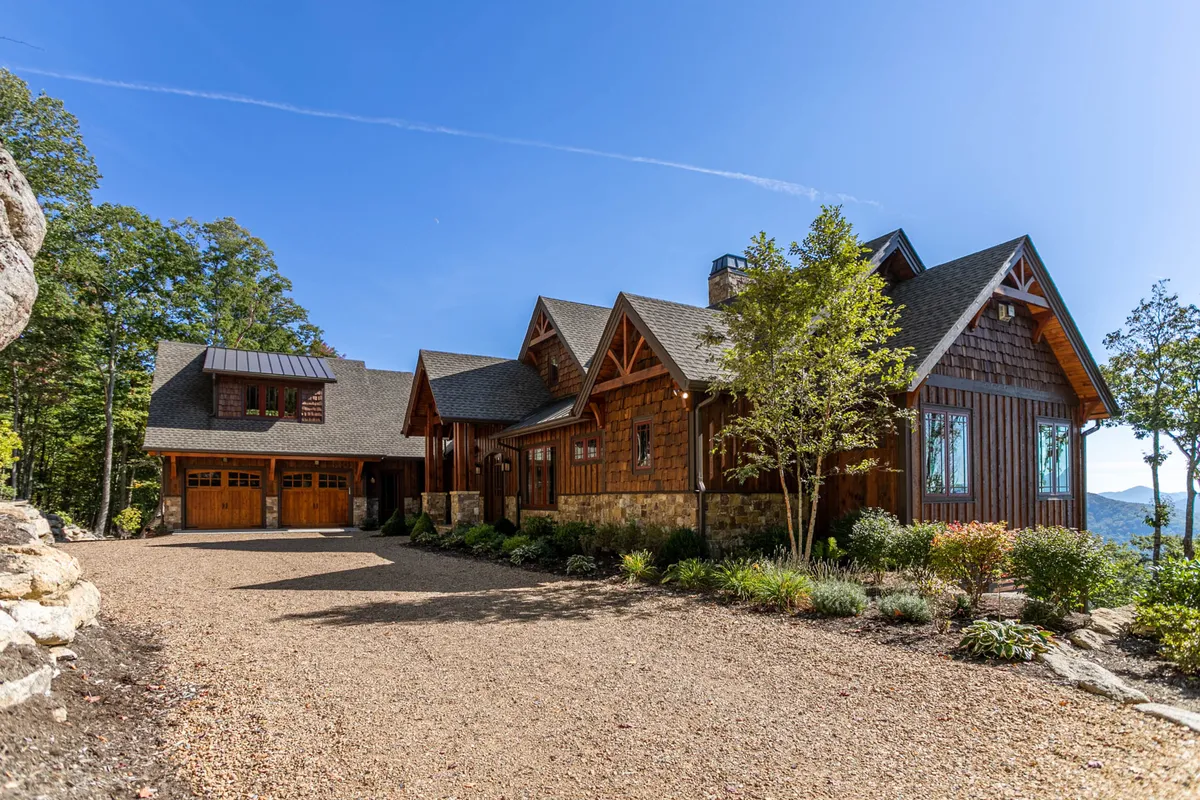 The hero image showcases the rustic wooden façade of a spacious mountain home, set against a stunning backdrop of sweeping Blue Ridge Mountain vistas. A stone pathway winds through the lush, natural landscape, inviting guests toward the home. The inviting outdoor space hints at cozy fire pit evenings, outdoor dining, and family gatherings, all surrounded by serene nature.