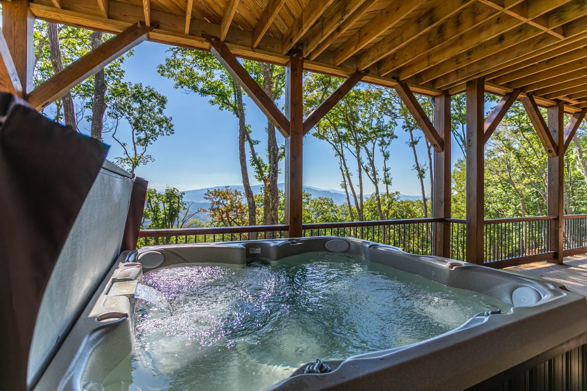 This exterior image centers on a luxurious outdoor hot tub set against a scenic mountain backdrop. Wooden beams frame the space, and the hot tub is positioned for maximum enjoyment of the panoramic view. The mood is tranquil, with hints of the sun setting over the mountains, inviting guests to relax and soak in the beauty and serenity of the location.