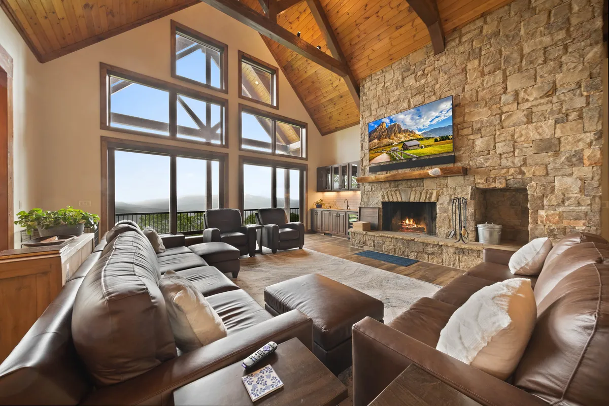 A spacious lounge with a large stone fireplace, comfortable seating area, and a television mounted above. Expansive windows offer views of the outdoors, blending cozy interior relaxation with the natural beauty beyond.